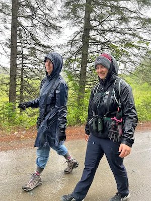Two young people (Fern and Maddie) walking on a wooded dirt road in the rain with binoculars and holding trash.